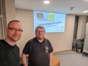 Michael und Hendrik schauen freundlich in die Kamera. Im Hintergrund die einzige Folie der Präsentation - die Begrüßung.